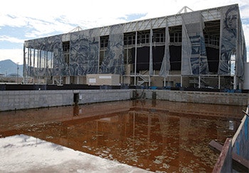 The Olympic practice pool has turned a nasty orange color in the months of neglect since the games last year. In the background, the mosaic exterior of the Aquatic Stadium hangs in tatters.