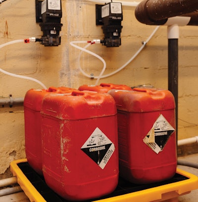 Acid carboys for managing pH in pool water sitting on a containment safety skid with acid chemical pumps in view.