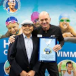 Swimming industry professionals posing with award at conference booth with swimmers in background