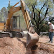 Yellow excavator digging on construction site with worker supervising near desert landscape and palm trees