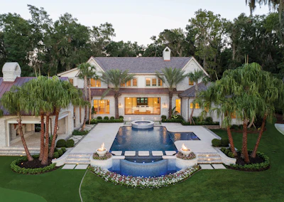 Luxury white modern mansion with illuminated windows, resort-style pool, spa, and palm tree landscaping at dusk