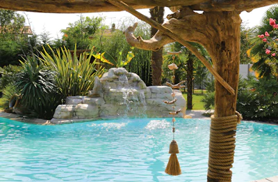 Tropical resort pool with stone waterfall feature, wooden pergola, and lush palm landscaping
