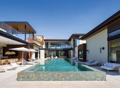 Modern luxury home with infinity pool, lounge seating, and multi-level architecture overlooking clear blue sky