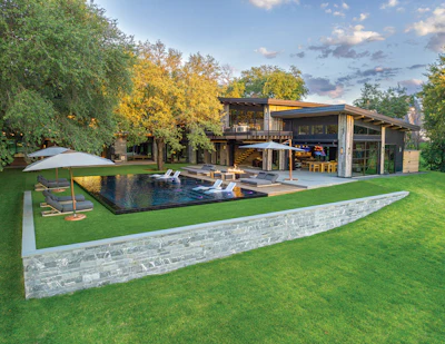 Modern residential home with infinity pool, blue umbrella, and manicured lawn surrounded by trees at dusk
