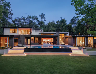 Modern architectural home with connected structures, illuminated windows, infinity pool, and manicured lawn at dusk