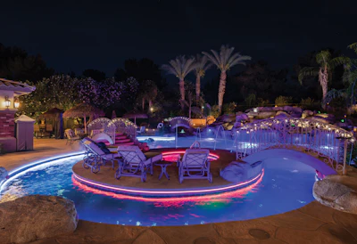 Luxury resort pool area illuminated at night with neon lighting, palm trees, and lounge seating