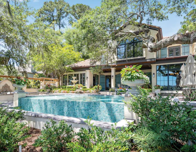 Modern luxury home with turquoise pool, stone patio, and mature trees in landscaped backyard