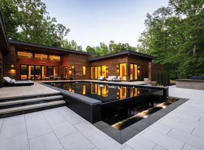 Modern residential home with illuminated interior, dark wood exterior, and reflecting pool at dusk surrounded by mature trees