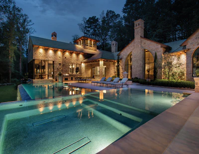 Luxury modern stone mansion with illuminated pool and lounge seating at dusk
