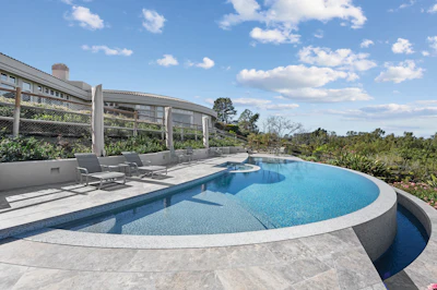 Modern curved swimming pool with blue water, lounge chairs, and contemporary residential architecture overlooking landscaped grounds