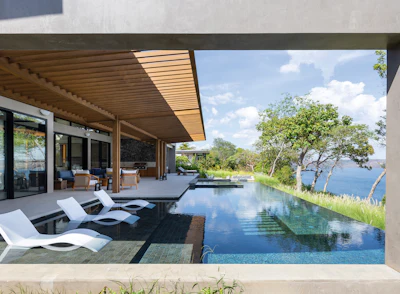 Modern luxury home with infinity pool overlooking scenic waterfront and tree-lined landscape under blue sky