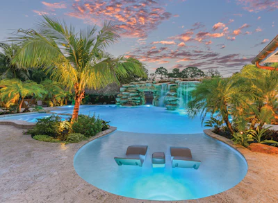 Luxury resort pool at sunset with turquoise water, palm trees, and waterfall feature under dramatic sky