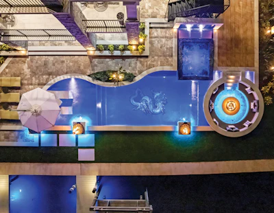 Luxury backyard pool and spa area with blue water, ambient lighting, lounge chairs, and landscaping at night