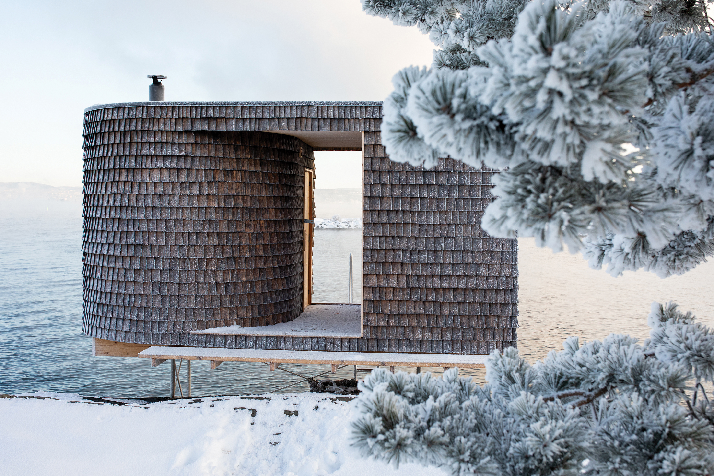 Modern wooden architectural structure with cylindrical tower and open frame overlooking snowy waterfront landscape