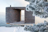 Modern wooden architectural structure with cylindrical tower and open frame overlooking snowy waterfront landscape