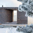 Modern wooden architectural structure with cylindrical tower and open frame overlooking snowy waterfront landscape