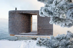 Modern wooden architectural structure with cylindrical tower and open frame overlooking snowy waterfront landscape
