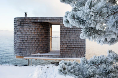 Modern wooden architectural structure with cylindrical tower and open frame overlooking snowy waterfront landscape