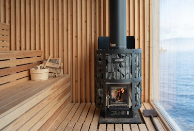 Modern sauna interior with black wood-burning stove, wooden benches, and ocean view through glass window