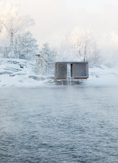 Minimalist wooden cabin on frozen lake surrounded by snow-covered trees in winter landscape