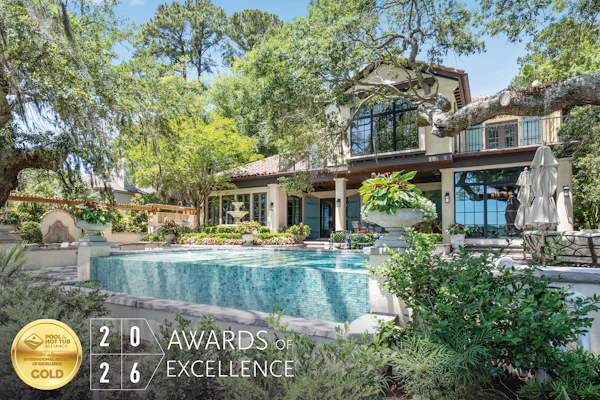 Luxury modern home with turquoise pool surrounded by mature trees and manicured landscaping, featuring 2026 Awards of Excellence gold badge