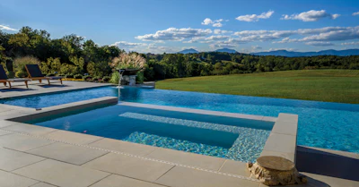 Luxury backyard with infinity pool, spa, lounge chairs overlooking green meadow and distant mountains under blue sky