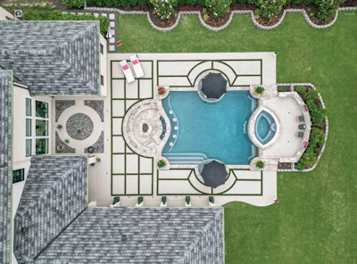 Aerial view of luxury home with resort-style pool, spa, and geometric patio design surrounded by manicured lawn