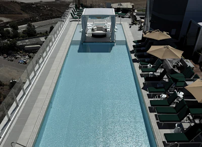 Aerial view of a modern rooftop pool with blue water, lounge chairs, tan umbrellas, and white railings