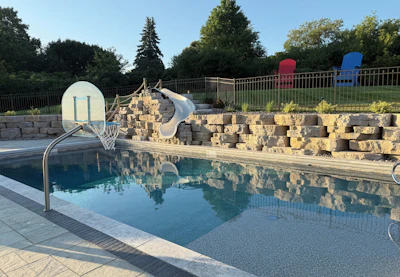 Backyard swimming pool with basketball hoop, stone retaining wall, and playground equipment in sunny residential setting
