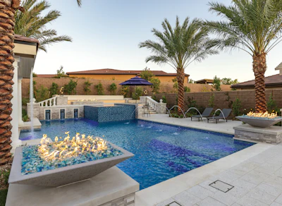 Luxury backyard with resort-style pool, fire pit, lounge chairs, palm trees, and brick walls at dusk