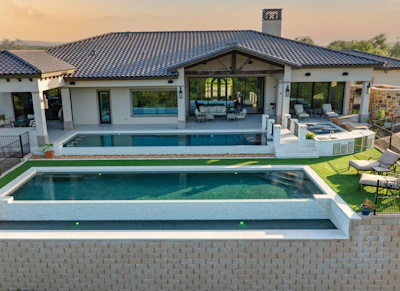 Modern luxury home with dark metal roof, white walls, infinity pool, spa, manicured lawn, and outdoor seating area at sunset