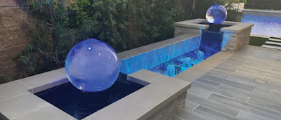 Modern backyard pool area with blue decorative spheres and illuminated water features surrounded by lush landscaping