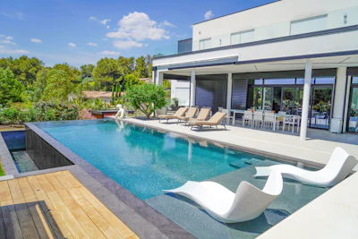 Modern luxury home with resort-style pool, lounge chairs, and manicured garden overlooking trees