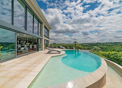 Modern luxury home with curved infinity pool overlooking green countryside landscape under blue sky