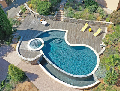 Aerial view of curved swimming pool with spa, wooden deck, yellow loungers, and lush landscaping
