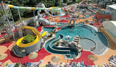 Aerial view of a colorful water park with turquoise pools, yellow water slides, and red safety surfaces surrounded by landscaping.