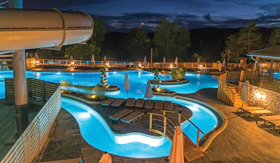 Illuminated resort pool complex at twilight with multiple pools, water slide, loungers, and forest backdrop