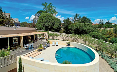 Modern home with curved pool, wooden deck, and Mediterranean landscape surrounded by olive trees and manicured gardens under blue sky