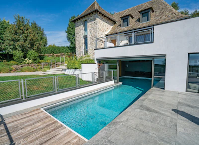 Modern two-story house with turquoise swimming pool, stone tower, white facade, and manicured lawn on sunny day