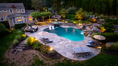 Aerial view of luxury backyard with illuminated swimming pool, stone patio, house, and landscaped gardens at dusk