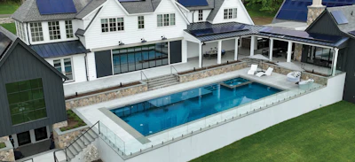Aerial view of modern luxury home with rectangular swimming pool, white exterior, and stone patio surrounded by manicured lawn.