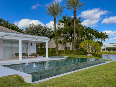 Modern luxury home with infinity pool, white patio, and tropical palm trees overlooking waterfront property