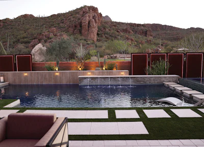 Modern desert resort pool with dark water, stone waterfall, rust-colored privacy screens, and native Arizona landscaping against rocky mountains.