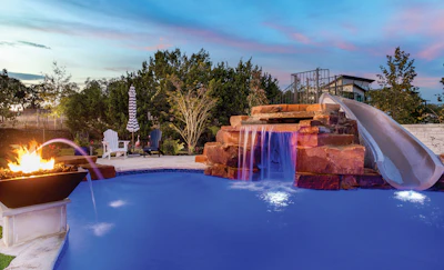 Luxury resort pool at dusk with illuminated waterfall, slide, fire pit, and scenic landscape backdrop