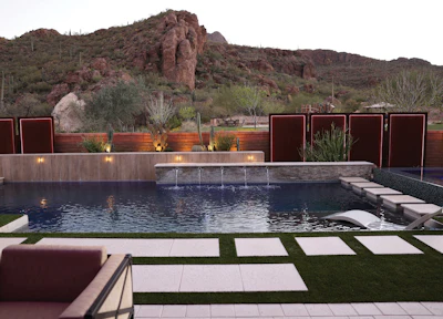 Modern luxury pool with dark water, stone waterfall feature, and desert mountain backdrop with red metal privacy screens and manicured lawn.