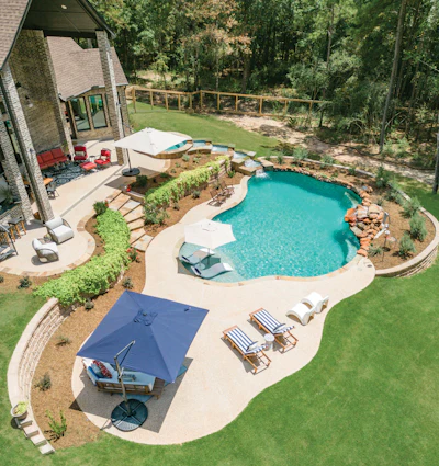 Aerial view of luxury backyard with turquoise swimming pool, patio furniture, umbrella, and manicured lawn surrounded by trees.