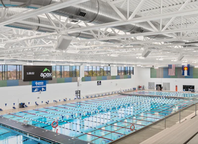 Modern indoor Olympic-sized swimming pool facility with multiple lap lanes, white steel ceiling structure, and APEX facility signage.
