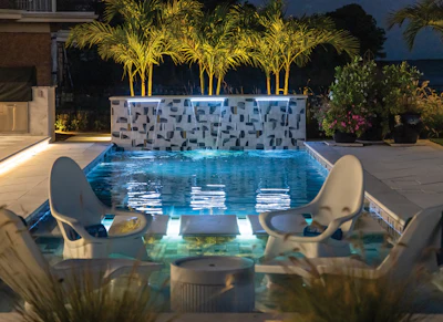 Illuminated luxury swimming pool at dusk with geometric wall, palm trees, and lounge chairs