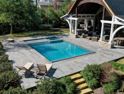 Luxury backyard with rectangular swimming pool, stone patio, lounge chairs, and covered pavilion with seating area surrounded by landscaping.
