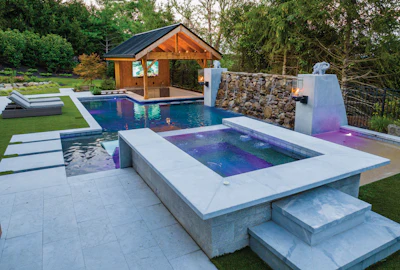 Luxury backyard pool complex with heated spa, wooden pool house, stone water feature, and manicured lawn.
